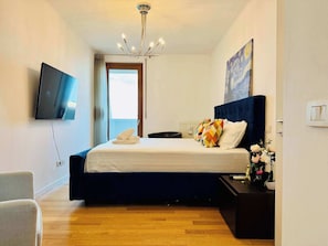 A bedroom with a bed, a TV, a chandelier, a painting, and a view through a window.