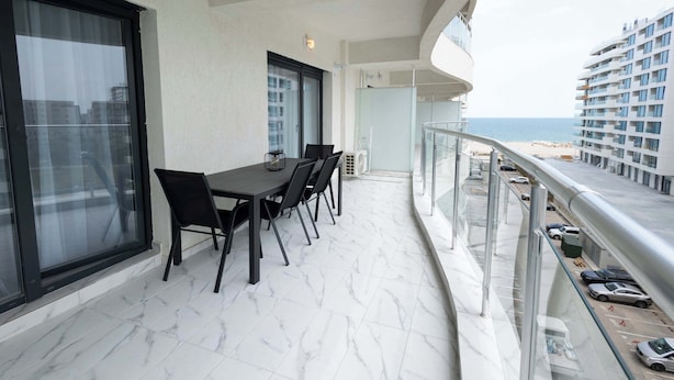 Un balcon avec une table et des chaises, une balustrade en verre, et une vue sur la plage et les bâtiments.