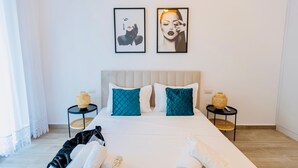 Een net opgemaakt bed met een witte deken, blauwgroene kussens en een zwarte plaids. Aan weerszijden van het bed staan twee kleine ronde tafels met gevlochten manden.