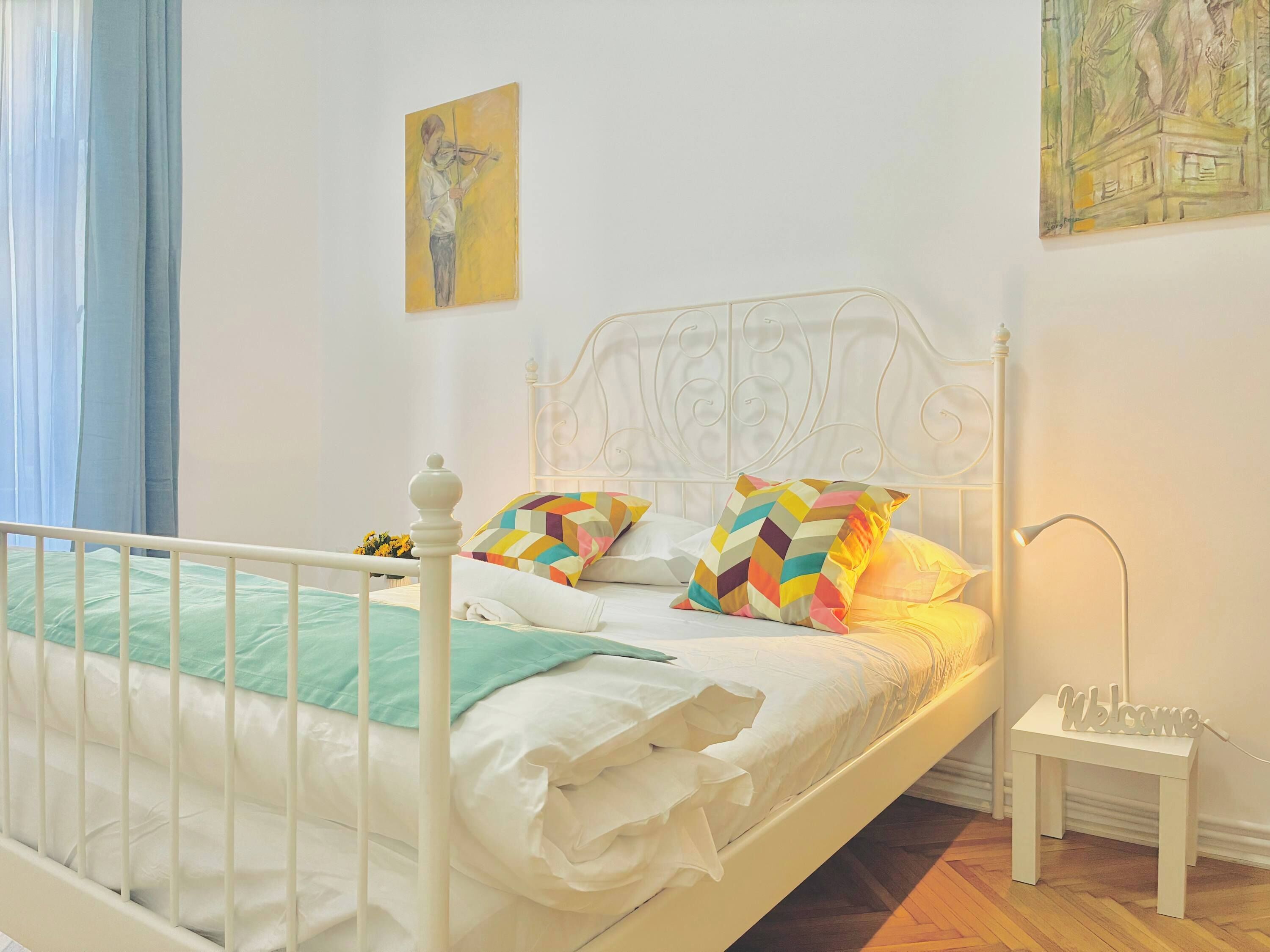 Una cama con estructura metálica blanca, un cojín colorido, una mesita de noche con lámpara y un cuadro en la pared.