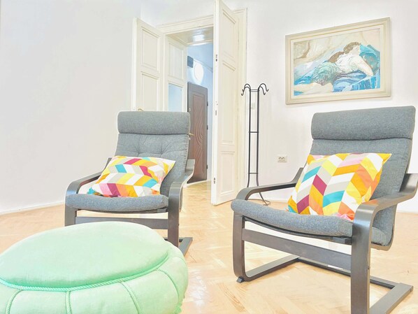Two grey armchairs with colorful geometric pillows, a green ottoman, and a painting on the wall.