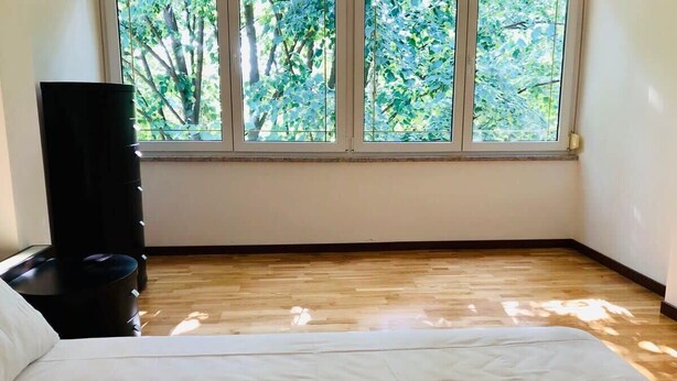 Une chambre d’hôtel avec une grande fenêtre, un plancher en bois, un lit avec du linge de lit blanc et un haut-parleur noir posé sur un support.