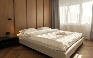 Een net opgemaakt bed in een kamer met houten paneelwanden en een groot raam met dunne gordijnen.