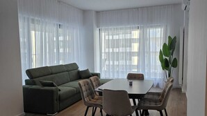 Moderni olohuone, jossa on vihreä sohva, ruokapöytä tuoleineen ja suuret ikkunat, joissa on läpikuultavat verhot.