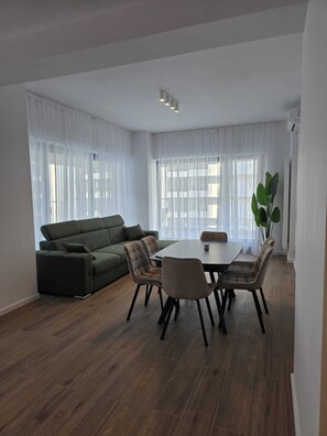 En moderne stue med en grøn sofa, et spisebord med stole og store vinduer med lette gardiner.