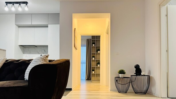 Un salon moderne avec un canapé marron foncé, un plancher en bois, un coin cuisine doté de meubles blancs et un couloir menant à une pièce avec une grande fenêtre et une plante.