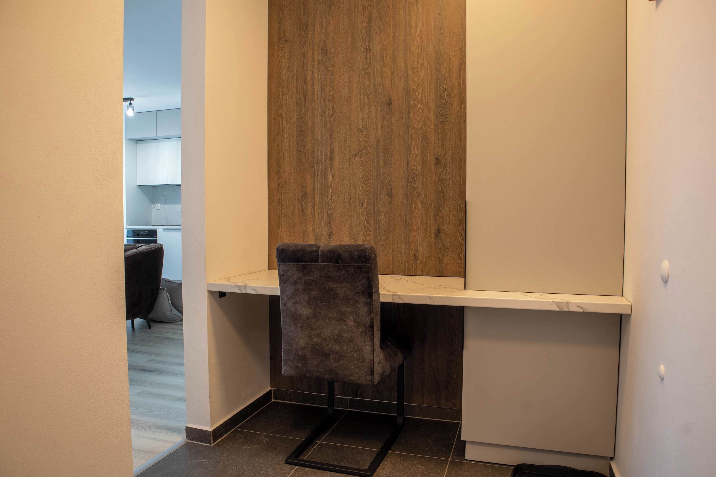 A small, modern kitchen with a wooden accent wall, a grey upholstered chair, and a white countertop.