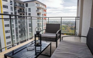 Een balkon met een glazen hekwerk, een rieten stoel, een met kussens uitgeruste bank, een glazen tafel en een kandelaar.