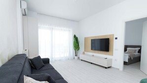 Un salon moderne avec un canapé gris, un téléviseur fixé au mur et une grande fenêtre ornée de rideaux légers.