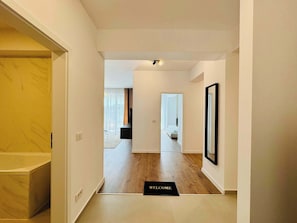 Un couloir doté d’un plancher en bois, d’un paillasson, d’un miroir à cadre noir et d’une porte donnant sur une chambre.