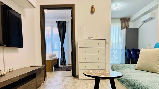 A modern living room with a flat-screen TV, a sofa, a small round table, a cabinet, and a bed with a window view. - Joy City Stay Mangalia 5 (Timișoara)