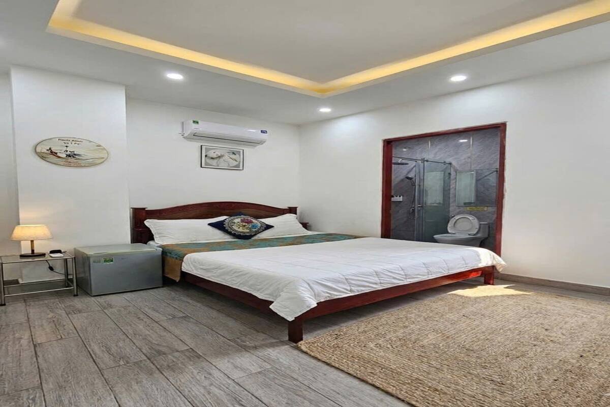 A bedroom with a bed, bedside table, lamp, air conditioning unit, and a bathroom with a toilet and mirror.