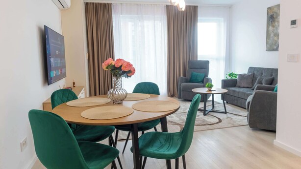 A modern living room with a dining area, grey sofas, a green armchair, a small table with a plant, and a painting on the wall.