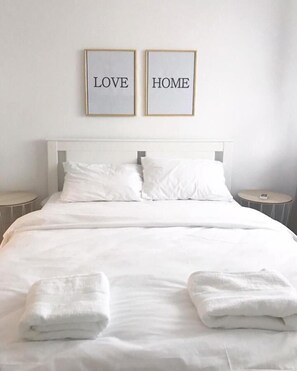 Ein ordentlich bezogenes Bett mit weißen Leinen und Kissen, zwei gerahmte Schilder an der Wand darüber mit den Aufschriften 'LOVE' und 'HOME', sowie zwei Nachttische mit Lampen.