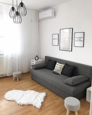 Ein modernes Wohnzimmer mit einem grauen Sofa, einem weißen, flauschigen Teppich, einem kleinen Hocker und einem Nachttisch mit einer Uhr.