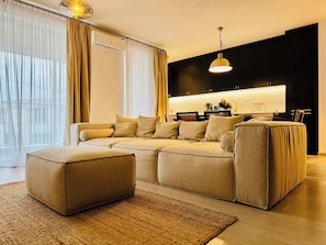 Een moderne woonkamer met een hoekbank, een salontafel en een keukenhoek met donkere kasten en een licht aanrecht.