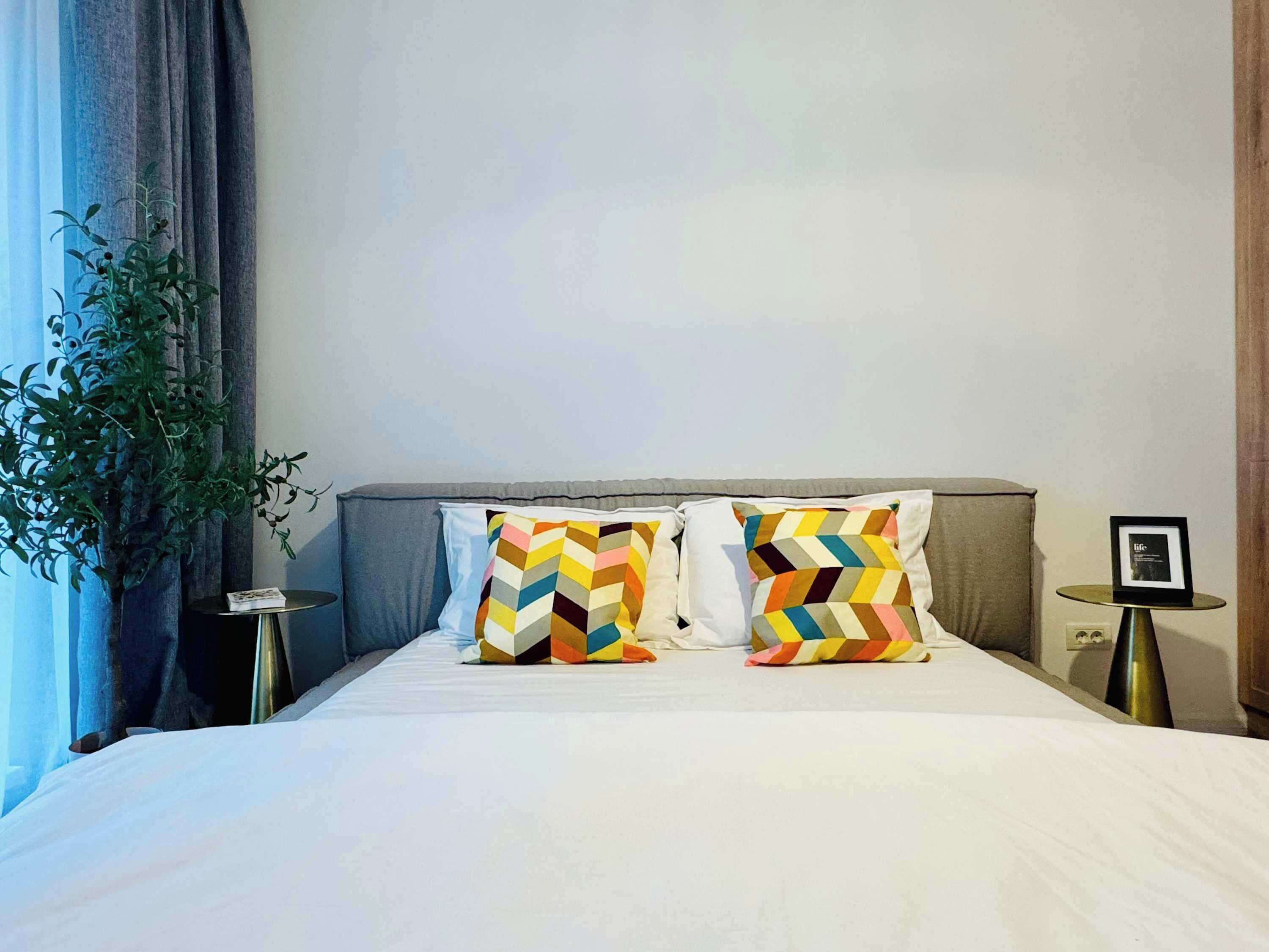 Una cama bien hecha con dos almohadones estampados, una mesita de noche con un marco de fotos y una planta en maceta junto a la ventana.