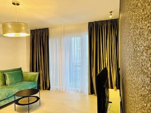 Ein Wohnzimmer mit einem grünen Sofa, einem runden Couchtisch, einem Flachbildfernseher und einem großen Fenster mit durchsichtigen Vorhängen.