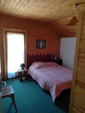 Une pièce comprenant un lit, une petite table, une lampe, un tableau au mur et une porte donnant sur l’extérieur.