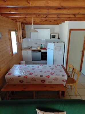 Une petite pièce agréable et chaleureuse, avec un plafond en bois, un lit recouvert d’une courtepointe à motifs floraux, une table à manger avec des chaises, un coin cuisine doté de meubles blancs et d’appareils électroménagers, et une fenêtre offrant une vue.