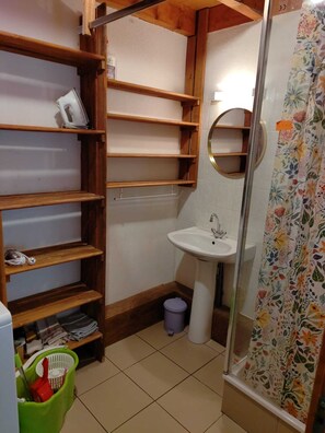 Un baño pequeño con un estante de madera, un lavabo, una cortina de ducha y un cubo de basura.