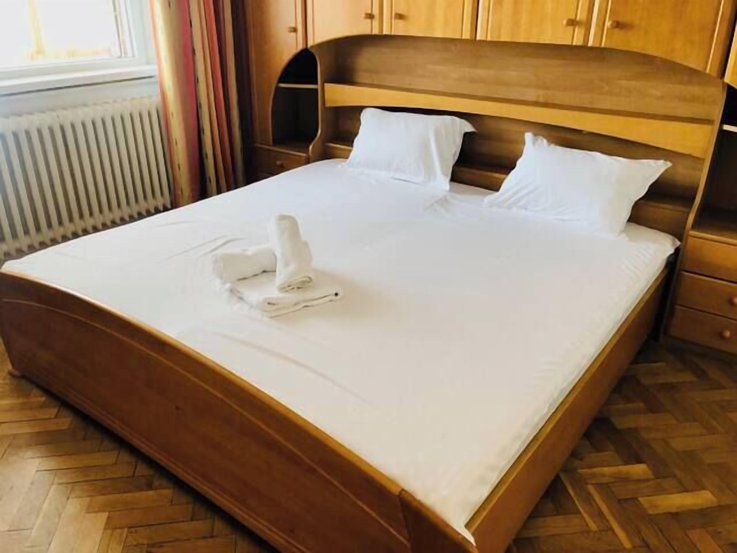 A neatly made bed with white linens and a folded towel, wooden headboard, and a wooden floor.
