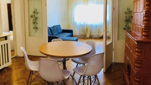 A dining area with a round table and four chairs, a blue sofa in the background, and a view of a balcony with a railing. - Joy City Stay Family Ultracentral (Timișoara)