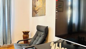 Ein modernes Wohnzimmer mit einem grauen Sessel, einem Holztisch mit einem 'Welcome'-Schild, einem Flachbildfernseher und einem Bild an der Wand.