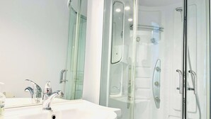 Ein modernes Badezimmer mit einem weißen Waschbecken, zwei Wasserhähnen, einer Glasdusche und einem Spiegel.