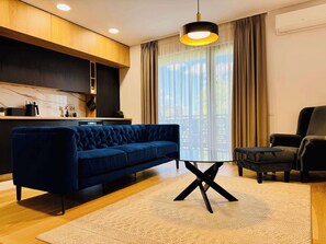 A modern living room with a blue sofa, a black armchair, a glass coffee table, wooden flooring, a beige rug, and a large window with curtains. - Joy City Stay Monarch 14 (Timișoara)