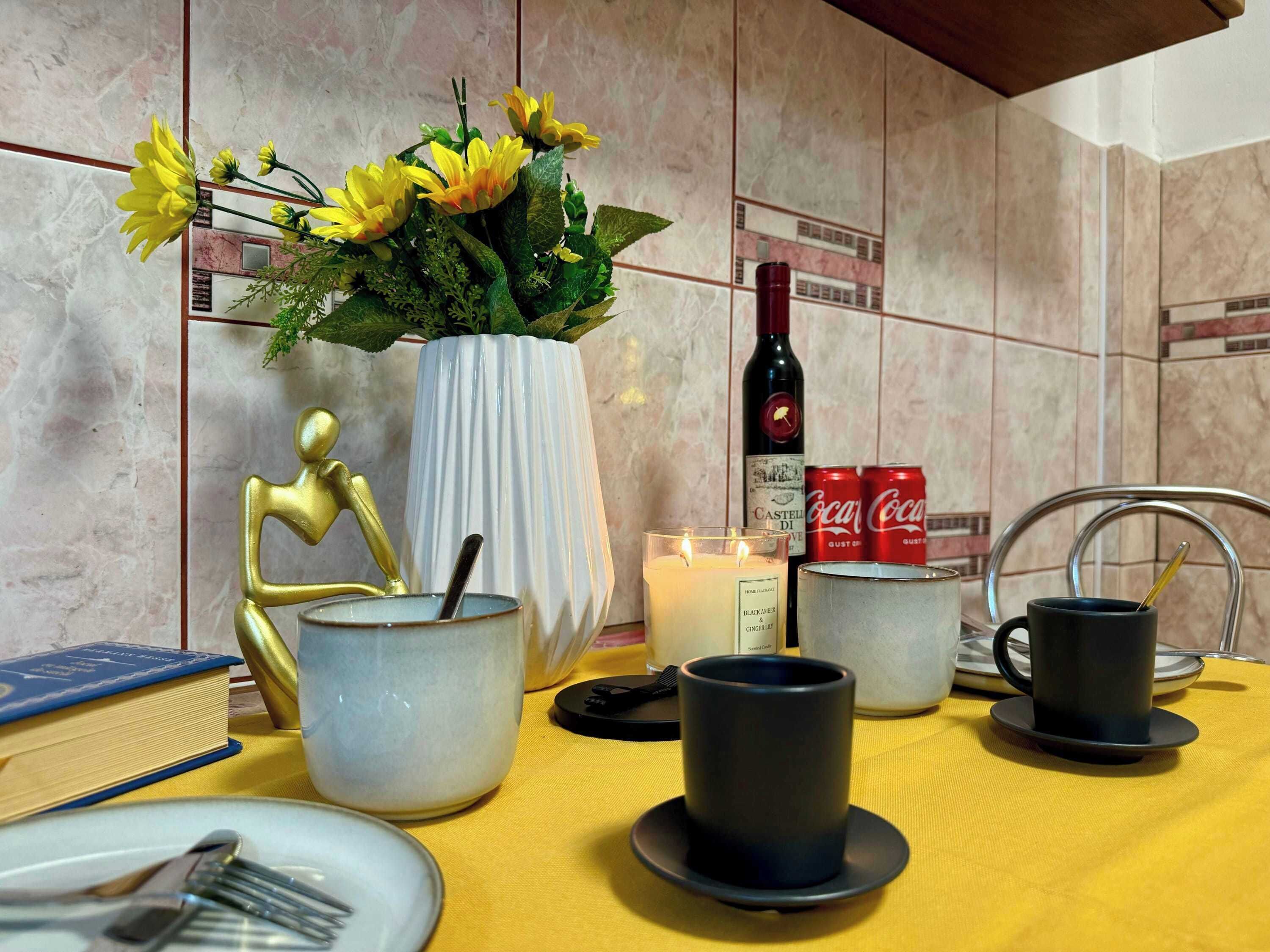 A dining table with a vase of yellow flowers, a candle, a bottle of wine, and two cans of Coca-Cola.