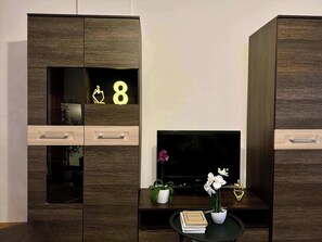 Una sala de estar moderna con un televisor de pantalla plana, armarios de madera, una pequeña mesa redonda con un libro y una planta en maceta, y objetos decorativos.