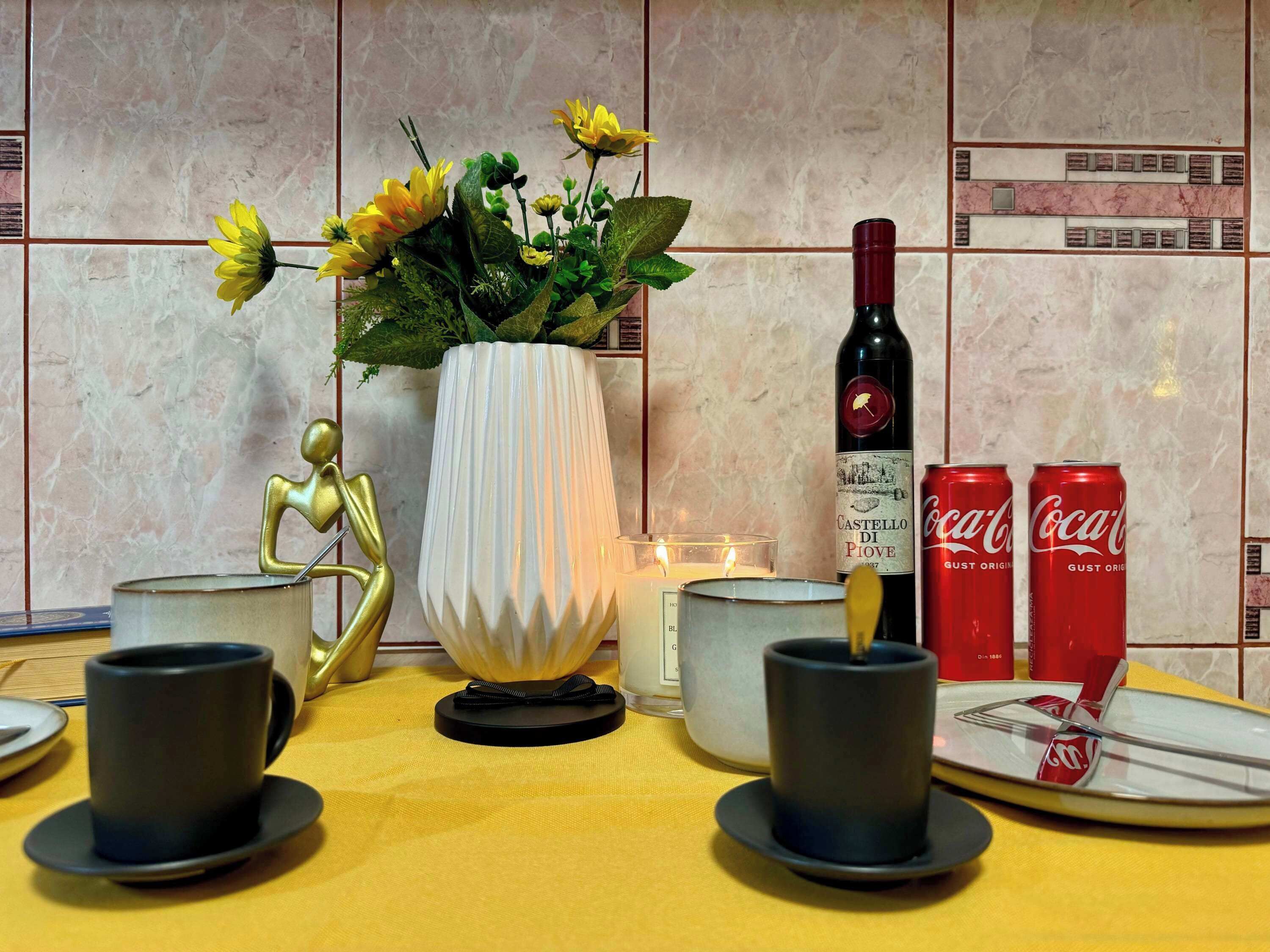 Ett matbord med en vas med blommor, en flaska vin, två burkar Coca-Cola och ett ljus.
