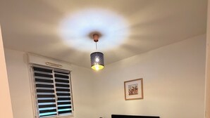 Un dormitori amb un llit, una tauleta de nit, una làmpada, una finestra amb persianes i un lluminari al sostre.