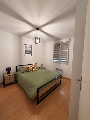 Ein Schlafzimmer mit einem Bett, einem Nachttisch, einer Lampe, einem Bild an der Wand und einem Fenster mit Jalousien.