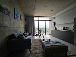 A modern living room with a sofa, a coffee table, and a dining area with chairs. There is a kitchen area with cabinets and a sink. The room has large windows providing a view of the city. - Studio Apartment in San Benito, with balcony & volcano views. (San Salvador)