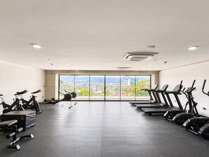 A gym with stationary bikes, treadmills, and weight machines, large windows with a city view. - Studio Apartment in San Benito, with balcony & volcano views. (San Salvador)