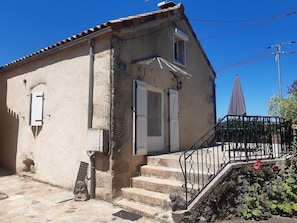 A building with a tiled roof, a staircase leading to a door, and a small garden area with a bench. - "Gîtes in la Couvertoirade "LE LARZAC (La Couvertoirade)