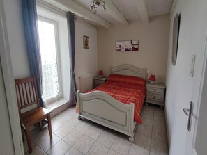 Un dormitorio con cama, mesitas de noche, una silla, un ventanal con cortinas y un radiador.