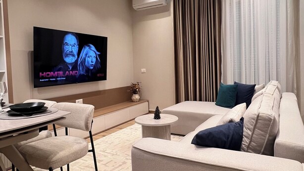 Een moderne woonkamer met een flatscreen televisie, een bank, een eethoek en een groot raam met gordijnen.