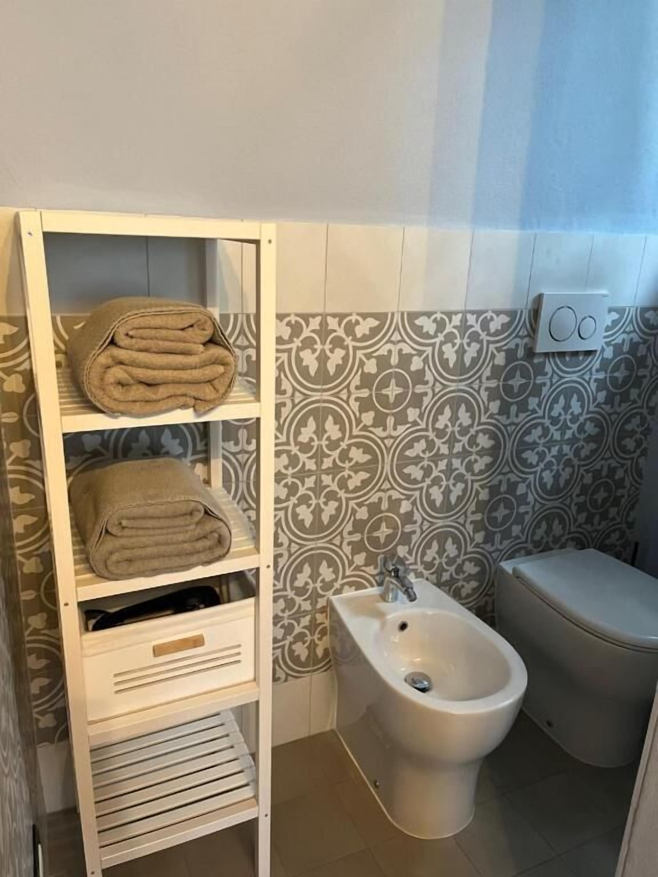 A bathroom with a bidet, a shelf with folded towels, and patterned wall tiles.