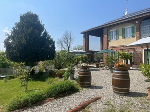 A courtyard with a large tree, outdoor seating, and a building with green shutters. - Noi Due B&B nel Monferrato (Quargnento)