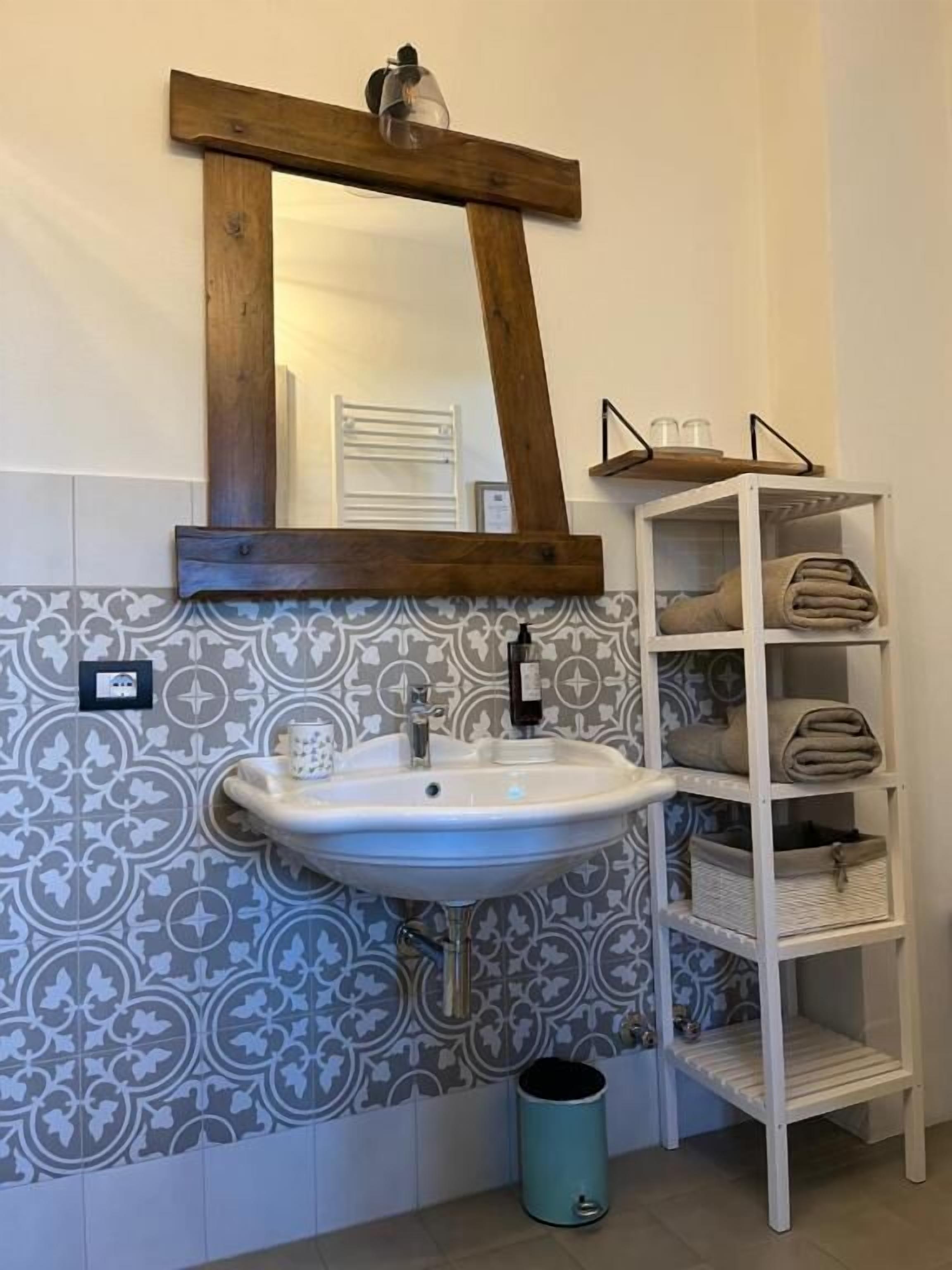 A bathroom with a white sink, a wooden-framed mirror, patterned tiles, a white shelf with towels, and a teal trash can.