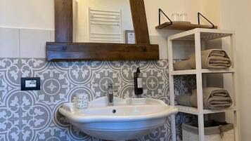 A bathroom with a white sink, a wooden-framed mirror, patterned tiles, a white shelf with towels, and a teal trash can.