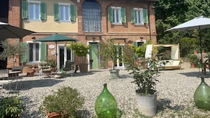 A two-story building with a brick facade, green shutters, and a central arched window. There are outdoor seating areas with tables and chairs, and potted plants are scattered around. - Noi Due B&B nel Monferrato (Quargnento)