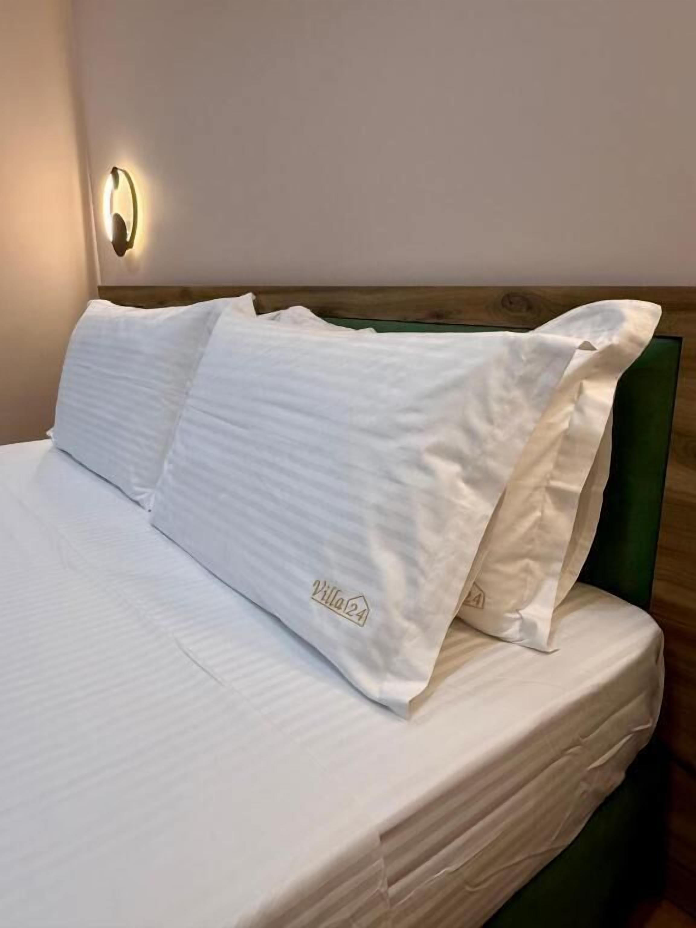 Una cama bien hecha con sábanas y almohadas blancas, un cabecero de madera y una lámpara de pared.