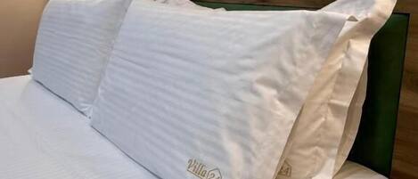 Una cama bien hecha con sábanas y almohadas blancas, un cabecero de madera y una lámpara de pared.