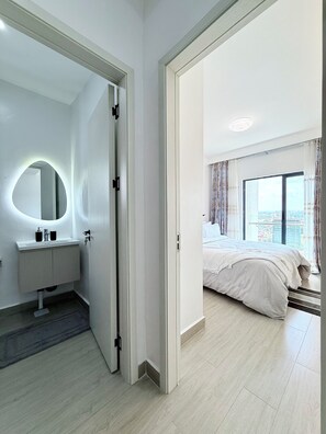 A modern bedroom with a large bed, a bathroom with a round mirror, and a view of the city through a large window.
