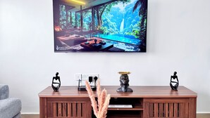 Una sala de estar moderna con un televisor grande que muestra un paisaje panorámico, una mesa consola de madera con objetos decorativos y un sofá gris.