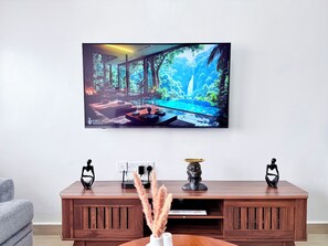Moderni olohuone, jossa on suuri televisio, jolla näkyy kaunis maisema, puinen konsolipöytä koriste-esineineen ja harmaa sohva.
